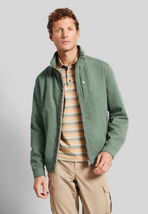 Green lightweight jacket with a high collar, zip closure, and side pockets, worn over a striped polo shirt. Plain beige cargo pants.