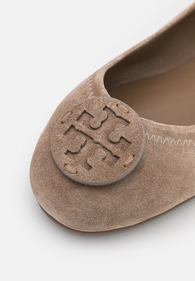 Suedes ballettleilighet i lys brun med et sirkulært, teksturert emblem på tåen. Overdelen har en glatt finish og subtile sømmer.