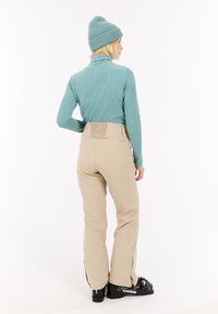 Un col roulé côtelé vert menthe, un pantalon beige avec des accents de fermetures éclair et des bottes de ski noires. Vue de dos, mettant en avant le design ajusté et les textures.