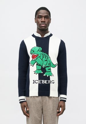 Mann trägt einen marineblau-weiß gestreiften Pullover mit einem großen grünen Dinosaurier und dem Schriftzug "ICEBERG" auf der Vorderseite, kombiniert mit beigen Hosen.