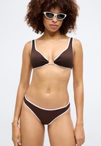 Ruda raukšlėta bikinis su baltais apvadais, turintis V formos viršūnę su priekiu detalėmis ir atitinkamomis apatinėmis dalimis. Modelis dėvi kačių akių saulės akinius.
