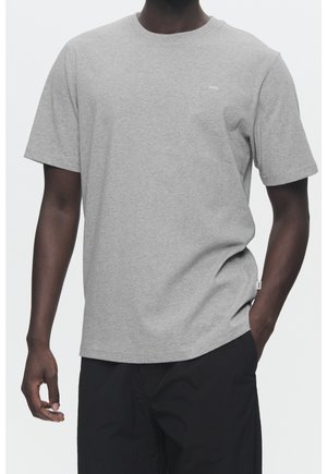 Mand iført en ensfarvet lysegrå kortærmet T-shirt og sorte bukser, stående med den ene hånd i lommen mod en hvid baggrund.