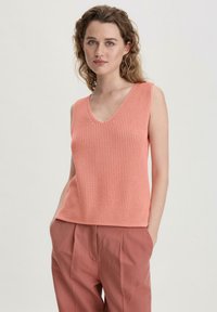 Helles rosafarbenes, gestricktes ärmelloses Oberteil mit V-Ausschnitt, das ein strukturiertes Muster und einen lässigen Schnitt aufweist, kombiniert mit lockeren, hellbraunen Hosen.