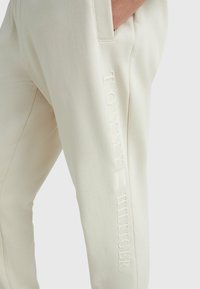 Pantalones de chándal beige confeccionados en un tejido suave, con un sutil logotipo en relieve "TOMMY HILFIGER" a lo largo del lateral como detalle de marca.