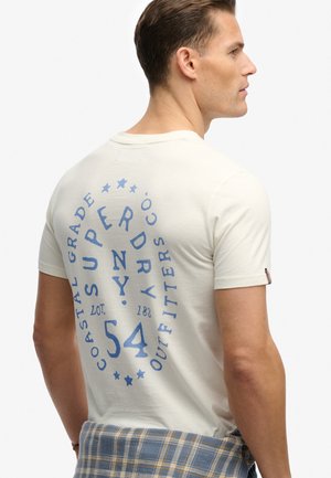 Homme portant un t-shirt Superdry couleur crème avec un logo texte bleu au dos, une chemise à carreaux claire nouée autour de la taille, vu de profil.