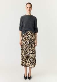 Vrouw die een donkergrijze trui draagt, midi rok met luipaardprint, zwarte hakken en een zilveren armband, staand tegen een effen lichte achtergrond.