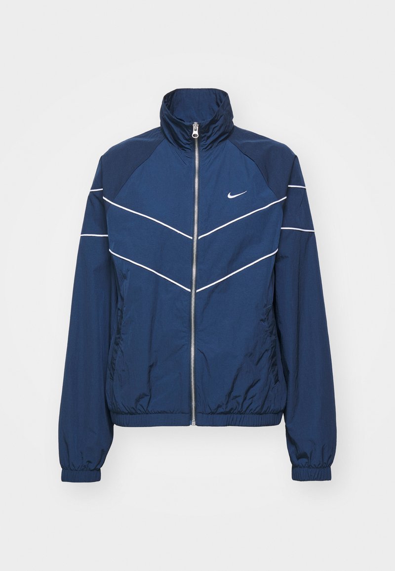 Veste coupe-vent marine avec un col haut, des rayures blanches accentuées, des poignets élastiques, une fermeture éclair à l'avant et un logo Nike sur la poitrine gauche.