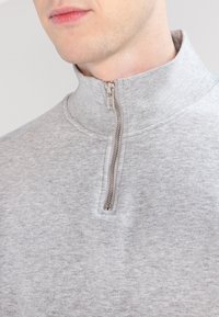 Grå zip-up sweatshirt med hög krage, med en metallisk dragkedja och ett texturerat tyg. Slät yta med subtila heatherade mönster.