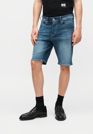 Mann trägt blaue Denim-Shorts, schwarze Socken, schwarze Leder-Loafer und ein schwarzes T-Shirt mit einer kleinen weißen Grafik.