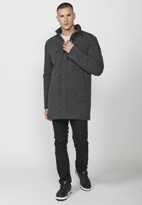 Abrigo largo gris oscuro con cuello alto, confeccionado en una tela texturizada, combinado con pantalones negros y zapatillas. Diseño simple y moderno.