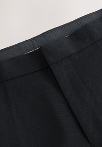 Gros plan sur un pantalon en tissu noir texturé avec une ceinture et une fermeture à crochet dissimulée sur un fond neutre.