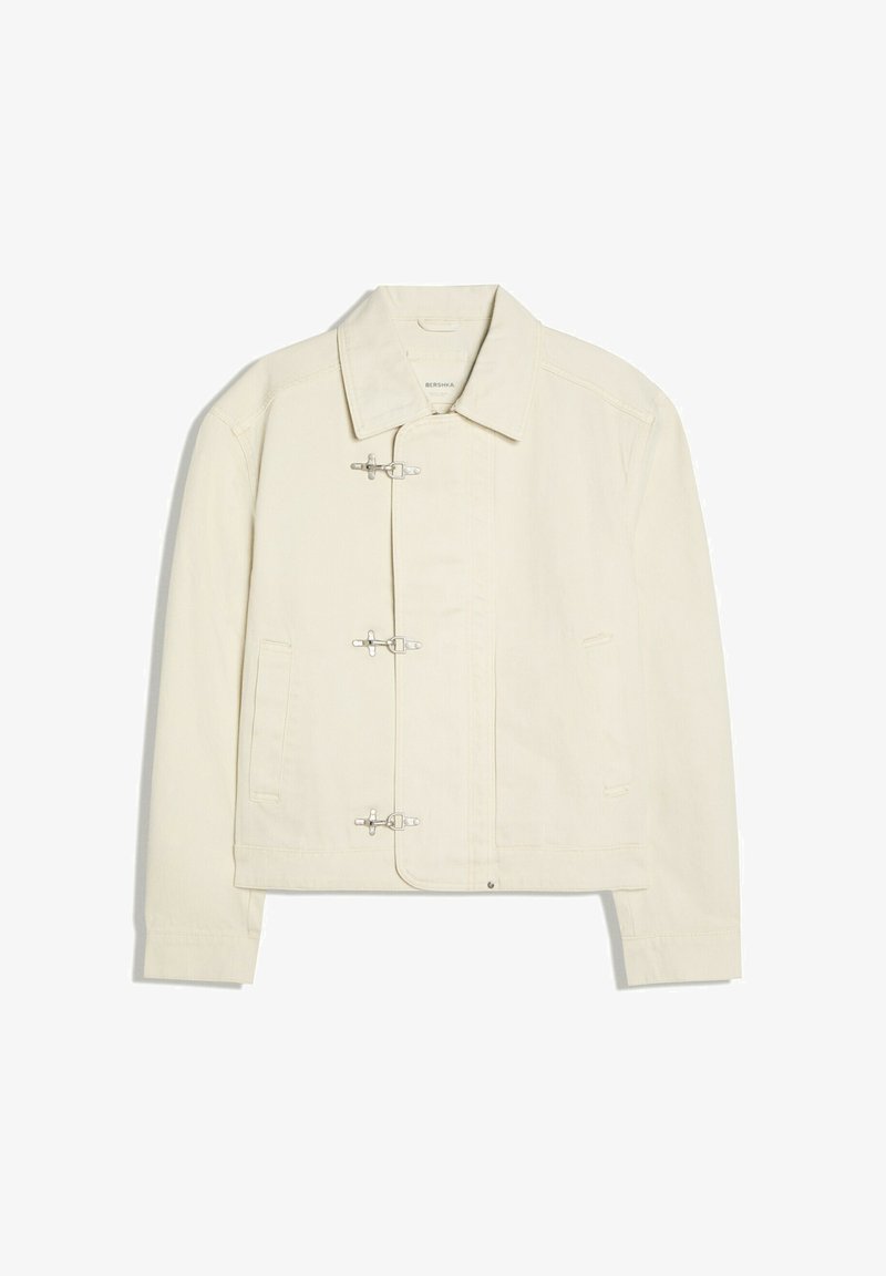 Veste blanc cassé avec un col et trois fermoirs à bascule argentés sur le devant, avec deux poches latérales et des manches longues.