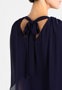 Blouse en soie bleu marine avec un design fluide, dos ouvert avec un détail de nœud, et texture lisse.