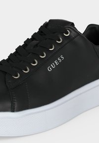 Μαύρο δερμάτινο αθλητικό παπούτσι με μαύρα κορδόνια, μεταλλικά ρουλεμάν, λευκό λογότυπο "GUESS" στο πλάι και χοντρή λευκή λαστιχένια σόλα.
