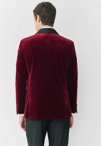 Piros bársonykabát fekete, kivágott gallérral, egy hátsó hasítékkal és három gombos mandzsettával. Sima textúra és testre szabott illeszkedés.