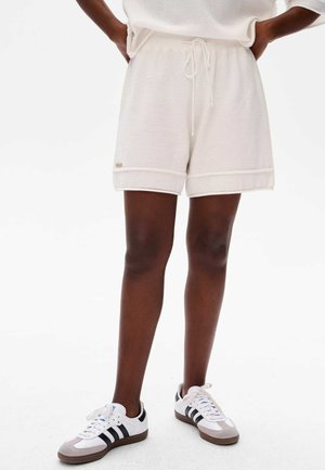 Shorts blancs en tricot avec une taille élastique et un cordon de serrage. Ourlets retroussés et un petit logo sur le côté. Portés avec des baskets noires et blanches.