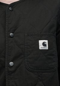 Giacca trapuntata nera con tasca applicata e logo bianco, chiusure con bottoni e finitura in tessuto strutturato. Design moderno e funzionale.