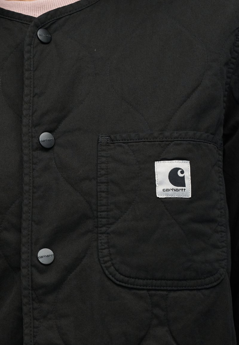 Zwarte gewatteerde jas met een opgestikte zak met een wit logo, knoopsluitingen en een geweven stofafwerking. Modern en functioneel ontwerp.