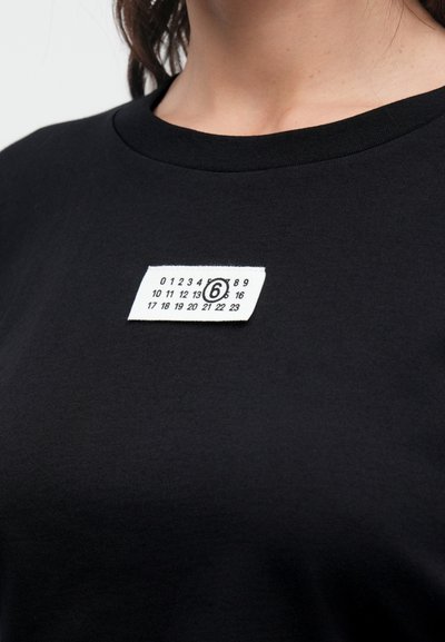 Schwarzes Baumwoll-T-Shirt mit einem weißen Etikett auf der linken Seite, auf dem Zahlen und die Zahl 6 in einem Kreis zu sehen sind.