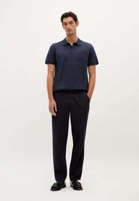 Dunkelblaues Poloshirt mit Brusttasche und kurzen Ärmeln, kombiniert mit lockeren dunklen Hosen und schwarzen Schuhen, vor einem schlichten hellen Hintergrund.