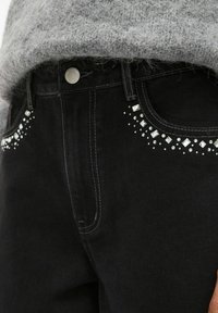 Crne traperice od denima s umjerenim strukom ukrašene dekorativnim rhinestones uz prednje džepove i suptilnom teksturom. Vidljiv detalj srebrnog dugmeta.