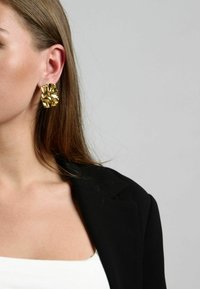 Pendientes de oro con un diseño orgánico y texturizado, que se asemeja a pliegues o ondas, llevados por una persona. Se puede ver un blazer negro y una blusa blanca.