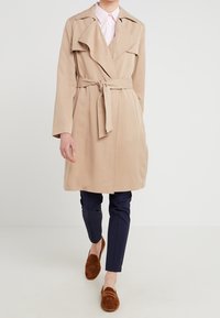 Beige trench coat med bälte i midjan, lagerkrage och långa ärmar, i kombination med marinblå byxor och bruna mockaloafers.