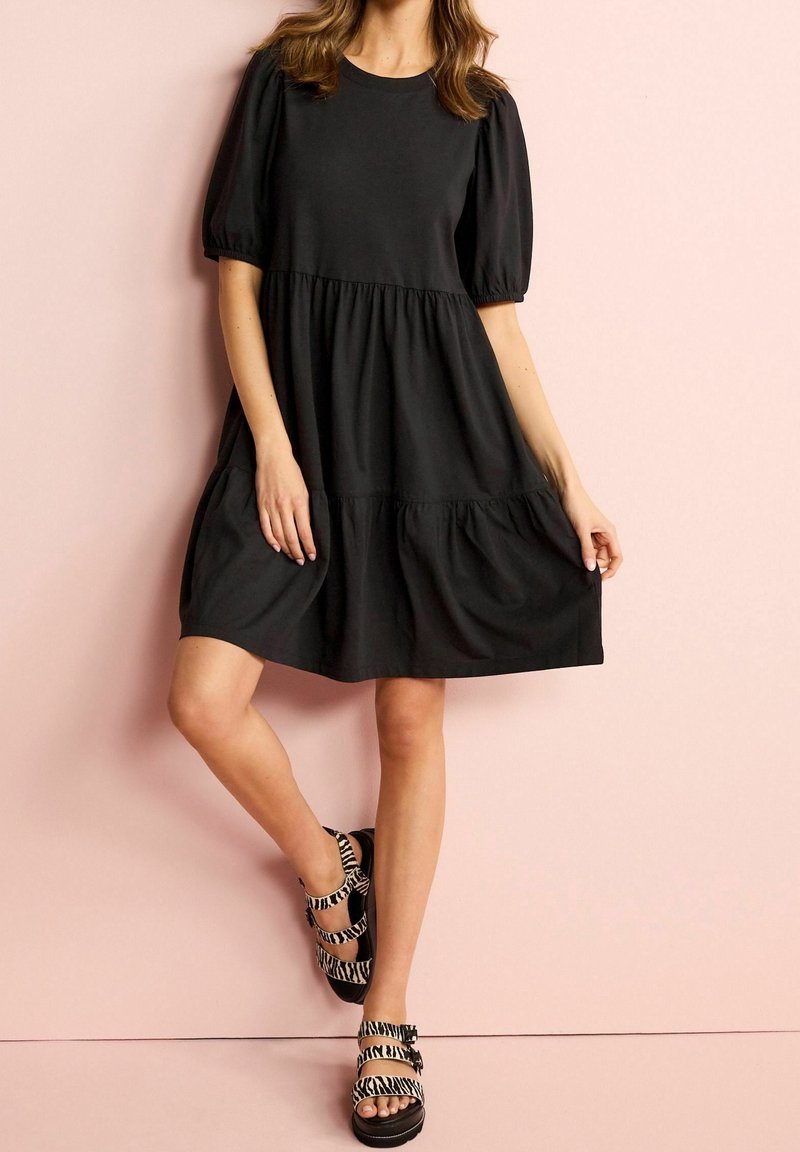 Femme portant une robe courte noire à manches bouffantes et des sandales à boucle imprimé zèbre, debout contre un mur rose clair.