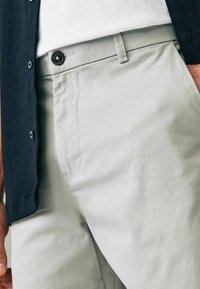 Hellgraue Hose mit glatter Textur, ausgestattet mit einer Vordertasche und einem schwarzen Knopfverschluss. Begleitet von einem dunkelblauen Hemd.