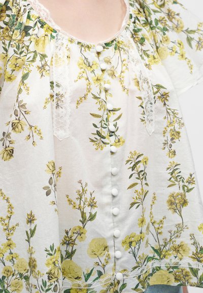 Blouse blanche avec imprimé floral jaune, boutons blancs verticaux et bordure en dentelle le long de l'encolure et des panneaux avant.