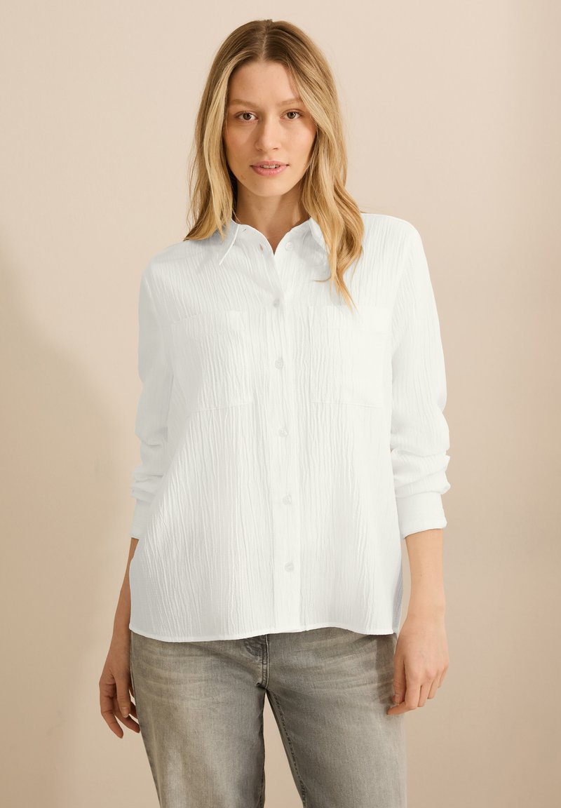 Hvid skjorte med knapper i struktureret stof, der har to frontlommer og lange, oprullede ærmer. Bæres med lysegrå jeans.