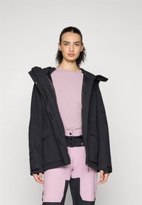 Melna izolēta jaka ar kapuci, kas aprīkota ar vairākiem kabatām un rāvējslēdzēju, uzlikta virs gaiši rozā krekla un rozā-pelēkām biksēm.