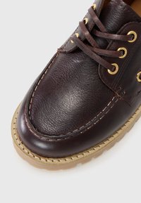 Brown leather shoe with a rounded toe, gold eyelets, and brown laces. Features a textured surface and a light-colored rubber sole.