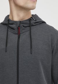 Grijze zip-up hoodie van een gestructureerde stof. Heeft een capuchon, zwarte koorden met rode accenten en een klein logo etiket op de kraag.