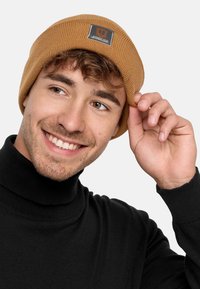 Tan geribbelde beanie met een logo patch, gedragen door een persoon met halflang krullend haar. Zwarte coltrui zichtbaar in het onderste deel van de afbeelding.