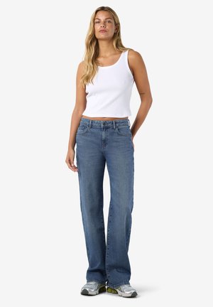 Jonge vrouw met lang blond haar, die een wit mouwloos crop top draagt, blauwe recht gesneden spijkerbroek en zilveren sneakers, staand met ontspannen handen.