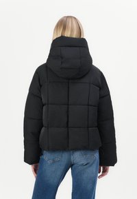 Chaqueta negra acolchada con capucha, que presenta un diseño acolchado y puños elásticos. El modelo lleva unos jeans azules, mostrando un corte ajustado y detalles mínimos.