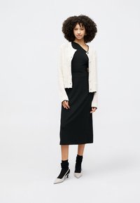 Juodas suknelė su prigludusia siluetu, derinama su kreminiu megztu kardiganu. Modelis vilki juodas raištines kojines ir smailėjantys basutės.