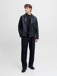 Mladý muž stojí, oblečený v čiernej koženej bunde, čiernom svetri, čiernych nohaviciach s prúžkami, čiernych topánkach, so strieborným retiazkou na krku, pozerá sa bokom.