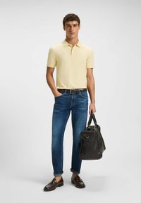 Man i gul pikétröja, blåjeans med uppvikta ben, bruna loafers, hållande en svart läderduffelväska, stående mot en neutral bakgrund.