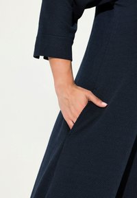 Námořnické modré šaty s texturovaným vzorem rybí kosti, tříčtvrtečními rukávy a postranními kapsami, které předvádějí uvolněný střih.