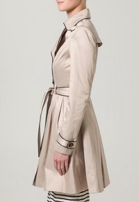 Beżowy trencz z dopasowaną talią, zapinanymi na guziki mankietami i paskiem do wiązania. Posiada kołnierz i czarne lamówki wzdłuż krawędzi. Gładka tekstura.