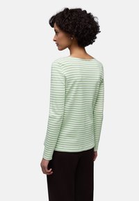 Mujer con cabello corto y rizado que lleva una camiseta de manga larga con rayas verdes claras y blancas, y pantalones negros, de perfil con la espalda visible.