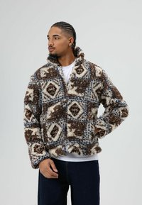 Wollen, meerkleurig jack in bruin, crème en blauw met een geometrisch patroon, hoge kraag en brede mouwen. Zachte textuur en losse pasvorm.