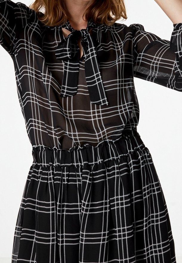 Femme portant un chemisier transparent à carreaux noir et blanc avec un col noué et une jupe taille haute assortie avec une ceinture élastique.
