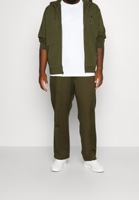 Olivgrön zip-up hoodie med en vit t-shirt under, ihop med olivfärgade joggerbyxor och vita sneakers. Slät tyg; avslappnad passform.