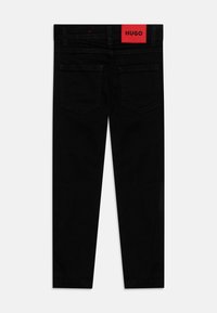 Jeans de mezclilla negra con corte ajustado. Presentan un parche rojo con el logo de HUGO en la cinturilla y dos bolsillos traseros con costuras decorativas.