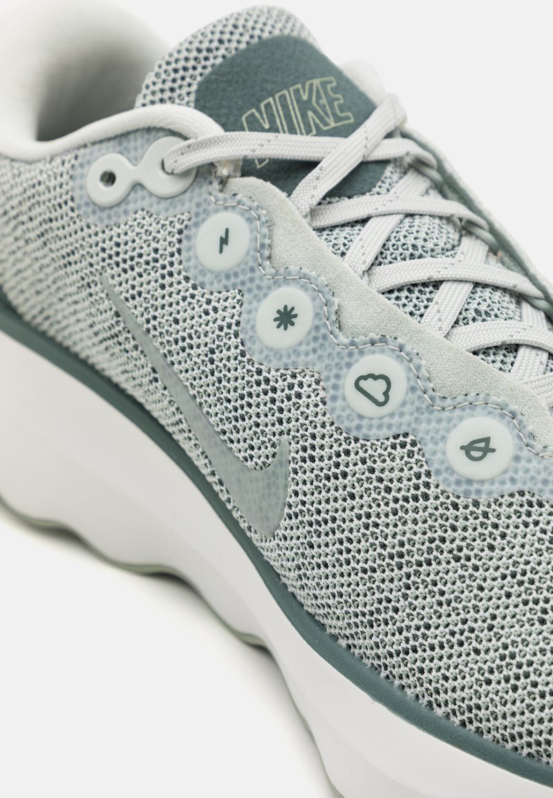 Nike Sneaker mit einem grauen Mesh-Obermaterial, texturierten Akzenten, weißer Sohle und markanten runden Knöpfen mit Symbolen und Logo auf der Zunge.