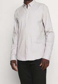 Lichtgrijs knoopshirt met lange mouwen, gemaakt van een textuurstof, voorzien van een kraag en witte knopen. G gedragen met zwarte broek.