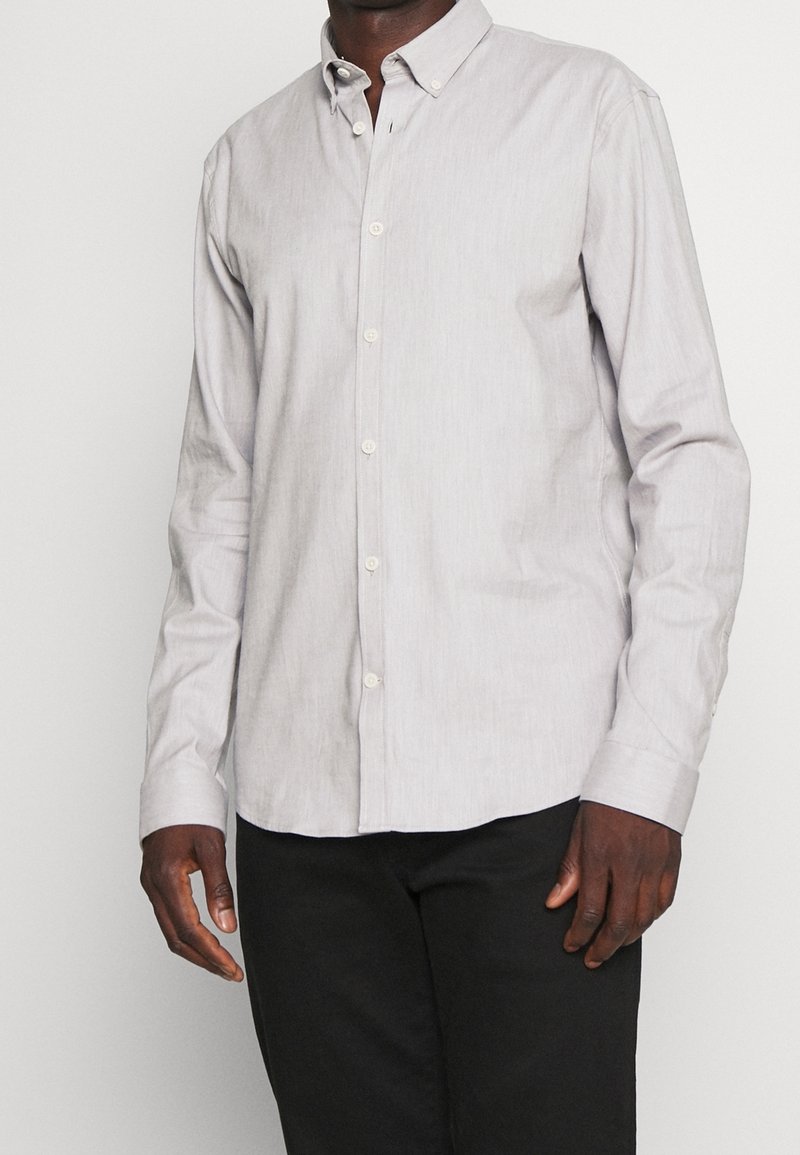 Lichtgrijs knoopshirt met lange mouwen, gemaakt van een textuurstof, voorzien van een kraag en witte knopen. G gedragen met zwarte broek.